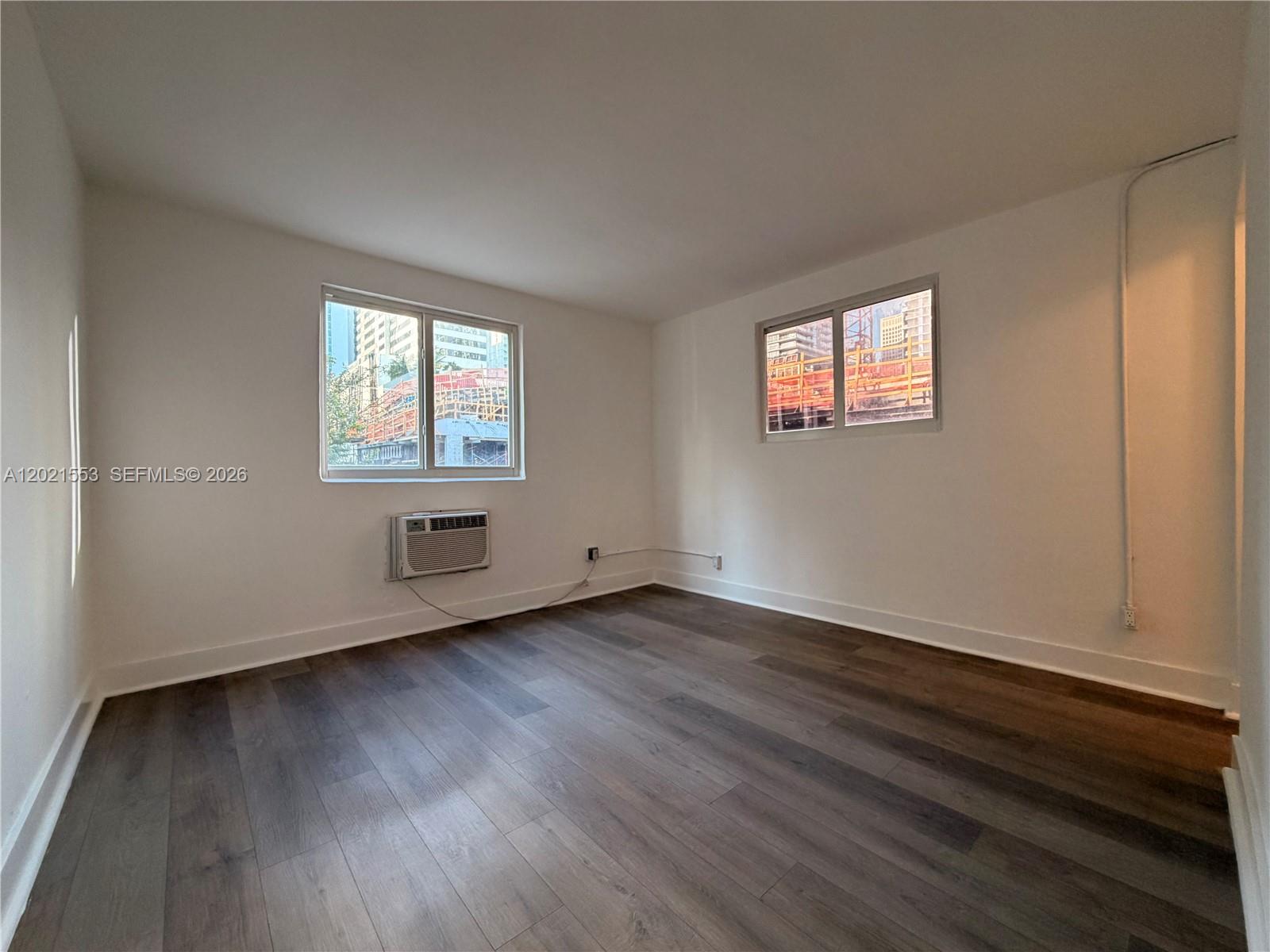 An empty room features white walls, dark wood-look flooring, two windows, and a wall-mounted air conditioner — 143 SW 9th St # 106, Miami FL 33130, MIAMI, FL