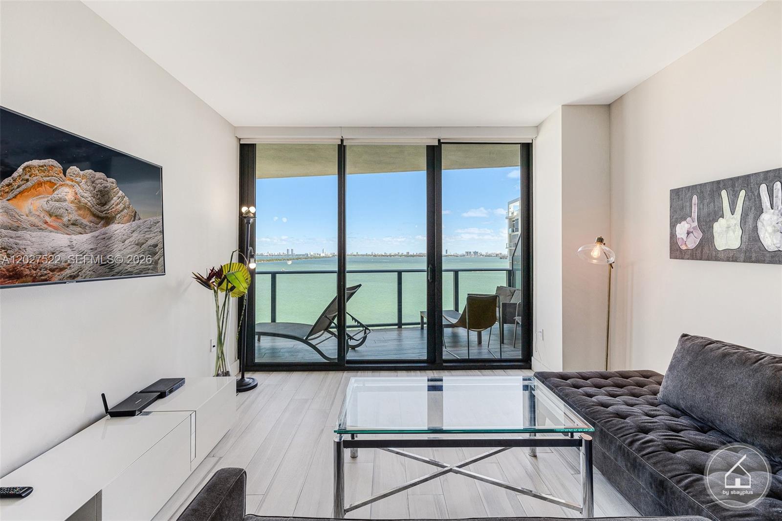 A modern living room features a glass coffee table, dark sofa, and balcony with water views — 460 NE 28th St # 1707, Miami FL 33137, MIAMI, FL
