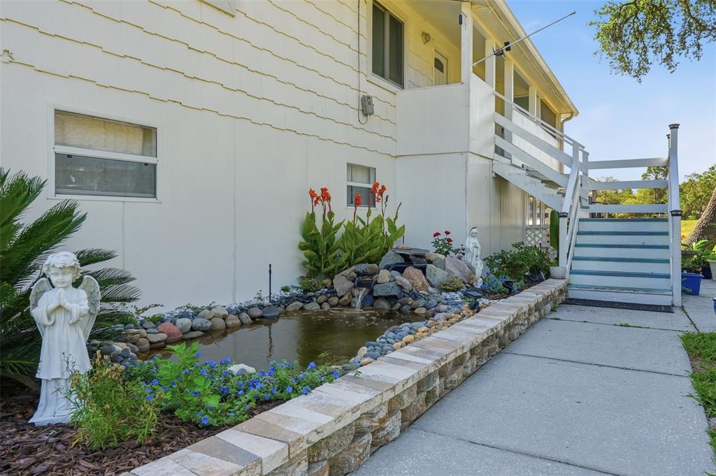 A small pond with a rock waterfall sits beside a house with an exterior staircase — 10226 LYNNHAVEN ROAD, SPRING HILL, FL
