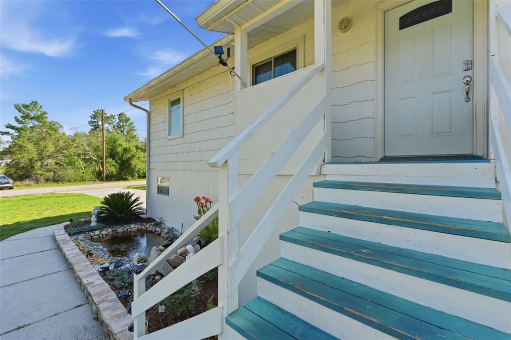 A white house exterior features a small pond, a walkway, and wooden stairs leading upward — 10226 LYNNHAVEN ROAD, SPRING HILL, FL