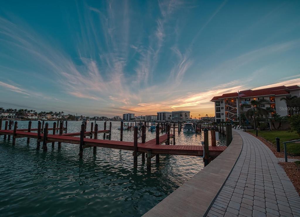 A paved walkway curves along a waterfront dock with boats and apartment buildings at sunset — 222 HARBOUR DRIVE UNIT 114, NAPLES, FL