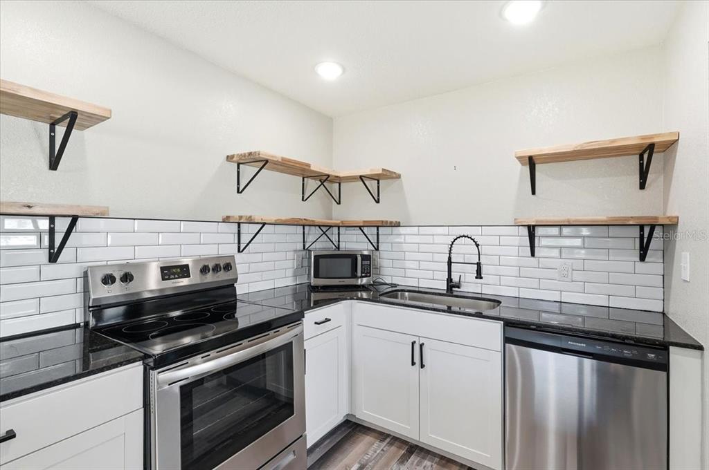A kitchen features white cabinets, black countertops, subway tile backsplash, and open wood shelving — 205 ANDOVER PLACE N UNIT 119, SUN CITY CENTER, FL