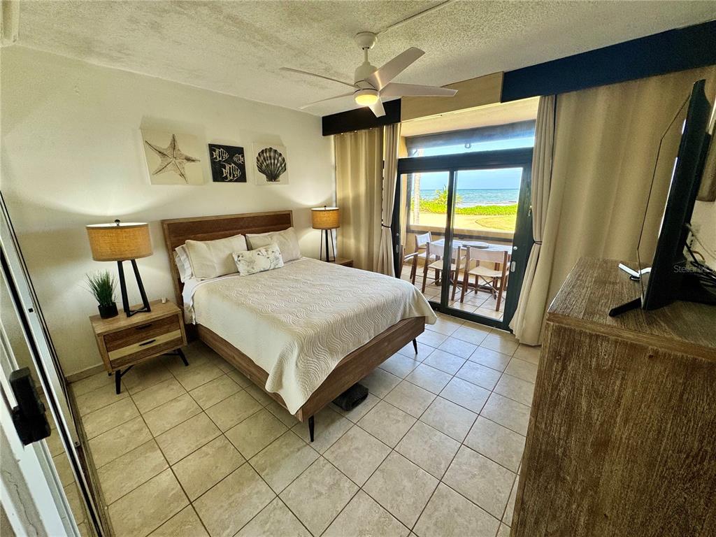 A bedroom with a queen bed, tiled floors, and a sliding glass door overlooking the ocean — 222 BEACH VILLAGE, HUMACAO, PR