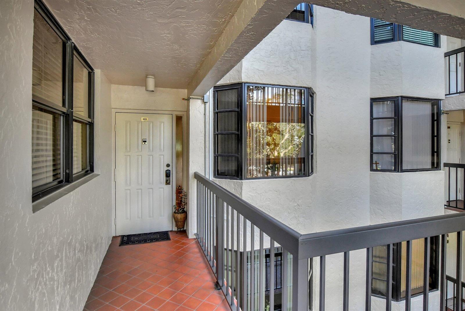 An exterior view of a third-floor apartment entrance with a tiled balcony and black railings — 6420 Boca Del Mar # 303, Boca Raton FL 33433, BOCA RATON, FL