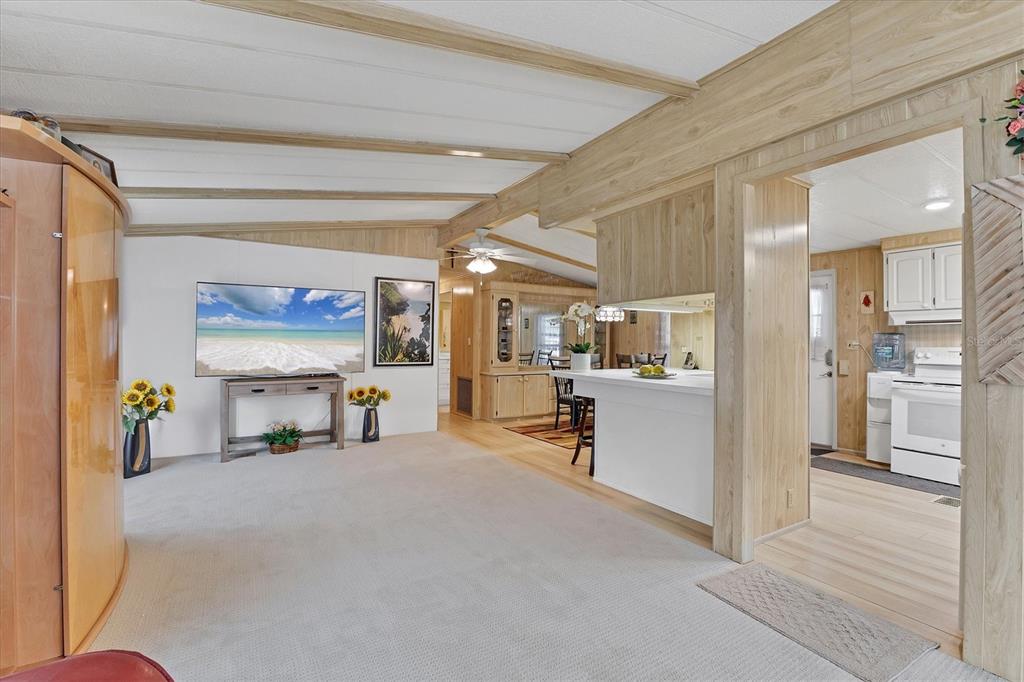 A living room with light carpet, wood-paneled walls, and a pass-through to the kitchen — 4532 12TH STREET COURT E, ELLENTON, FL