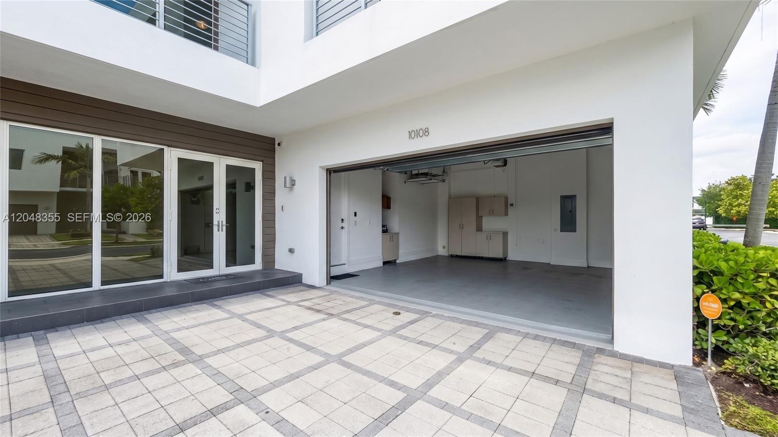 A modern home exterior features a paved courtyard, glass double doors, and an open garage — 10108 NW 77th St # 10108, Doral FL 33178, DORAL, FL