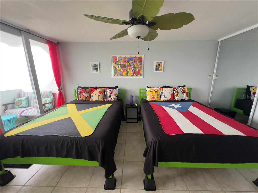 A bedroom features two beds with Jamaican and Puerto Rican flag blankets and a ceiling fan — Torre 1 ISLETA MARINA UNIT 10-R, FAJARDO, PR