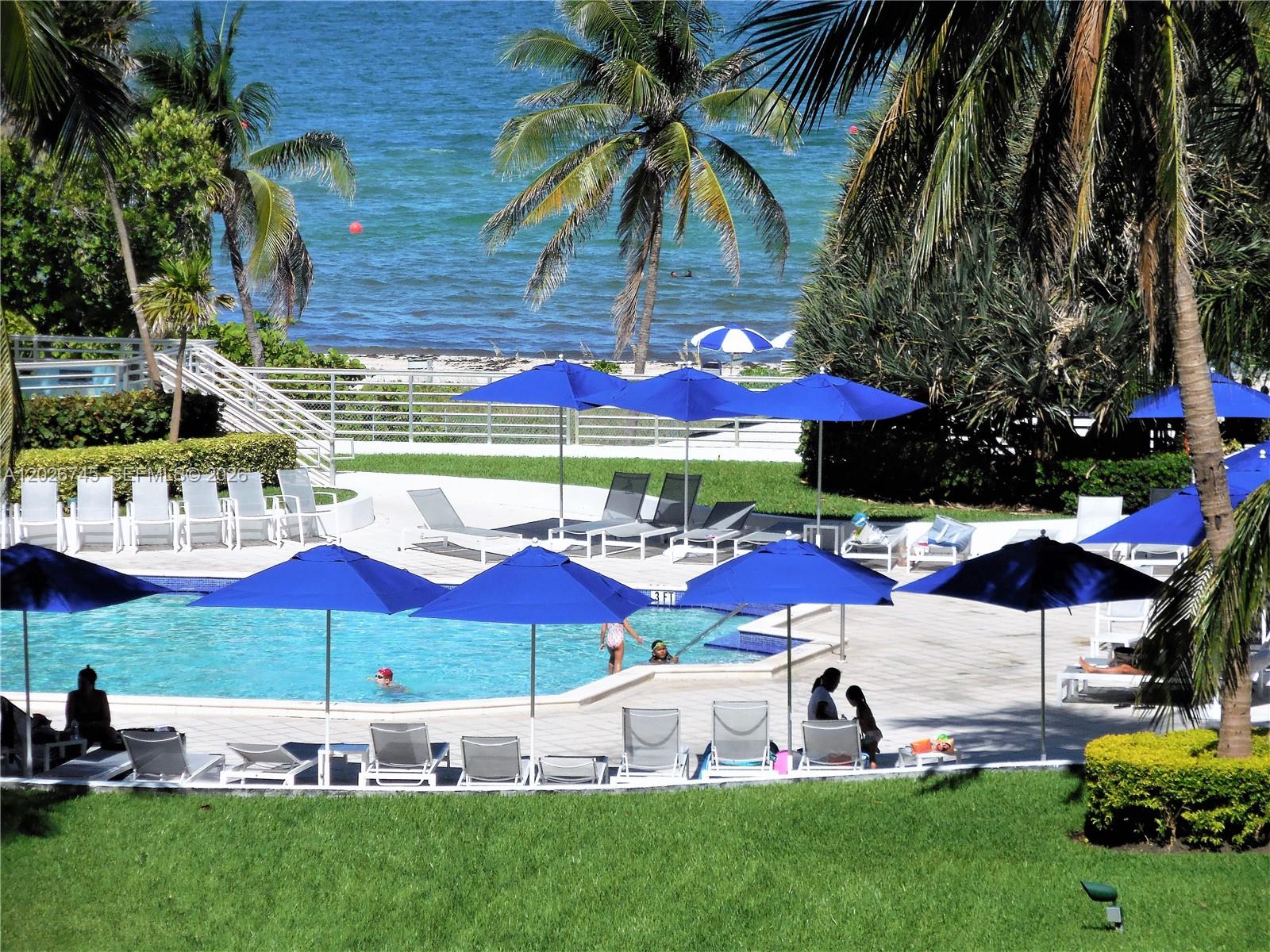 An outdoor swimming pool with blue umbrellas, lounge chairs, and a view of the ocean — 5151 Collins Ave # 519, Miami Beach FL 33140, MIAMI BEACH, FL