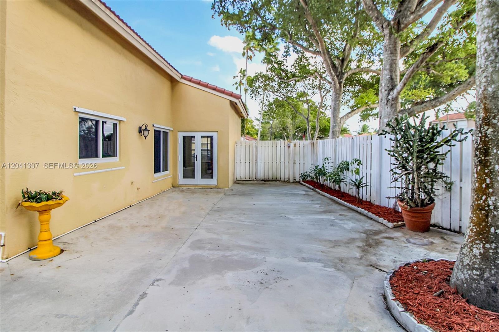 A concrete patio sits beside a yellow house with a white fence and trees — 12797 NW 13th St, Sunrise FL 33323, SUNRISE, FL