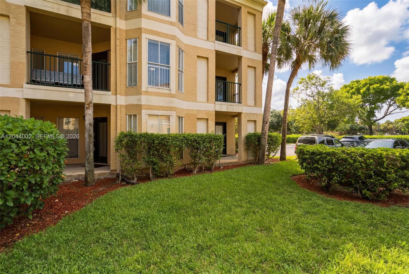 The exterior of a multi-story tan apartment building features balconies and a landscaped lawn — 977 Riverside Dr # 220, Coral Springs FL 33071, CORAL SPRINGS, FL