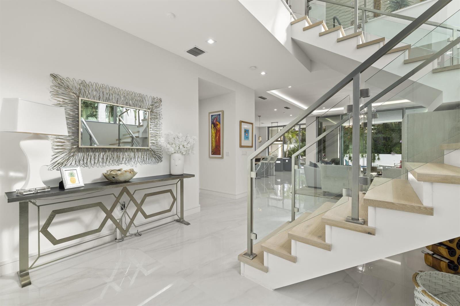 A modern foyer features a glass-railed staircase, marble flooring, and a decorative metal console table — 402 Ocean One Lane, Juno Beach FL 33408, JUNO BEACH, FL