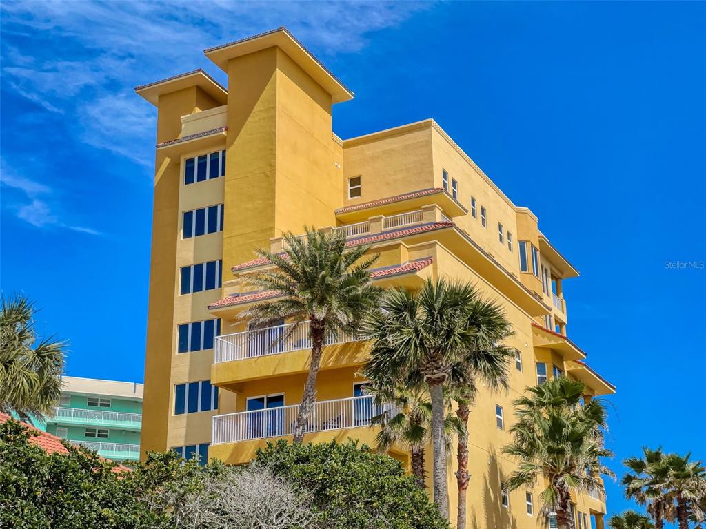 A multi-story yellow stucco condominium building features balconies and palm trees under a blue sky — 701 N ATLANTIC AVENUE UNIT 401, NEW SMYRNA BEACH, FL