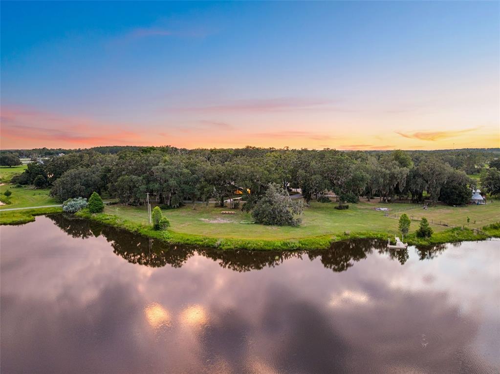 An aerial view shows a grassy yard with trees beside a lake at sunset — 33415 KIEFER ROAD, WESLEY CHAPEL, FL