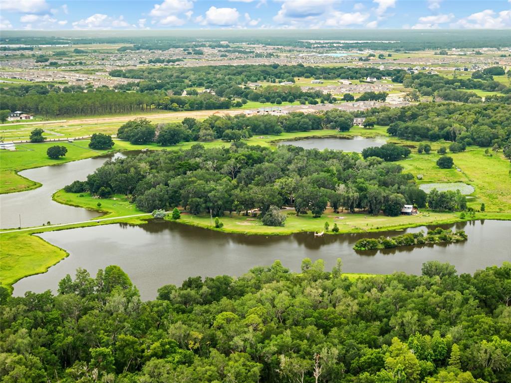 An aerial view shows a wooded island surrounded by a lake and grassy fields — 33415 KIEFER ROAD, WESLEY CHAPEL, FL