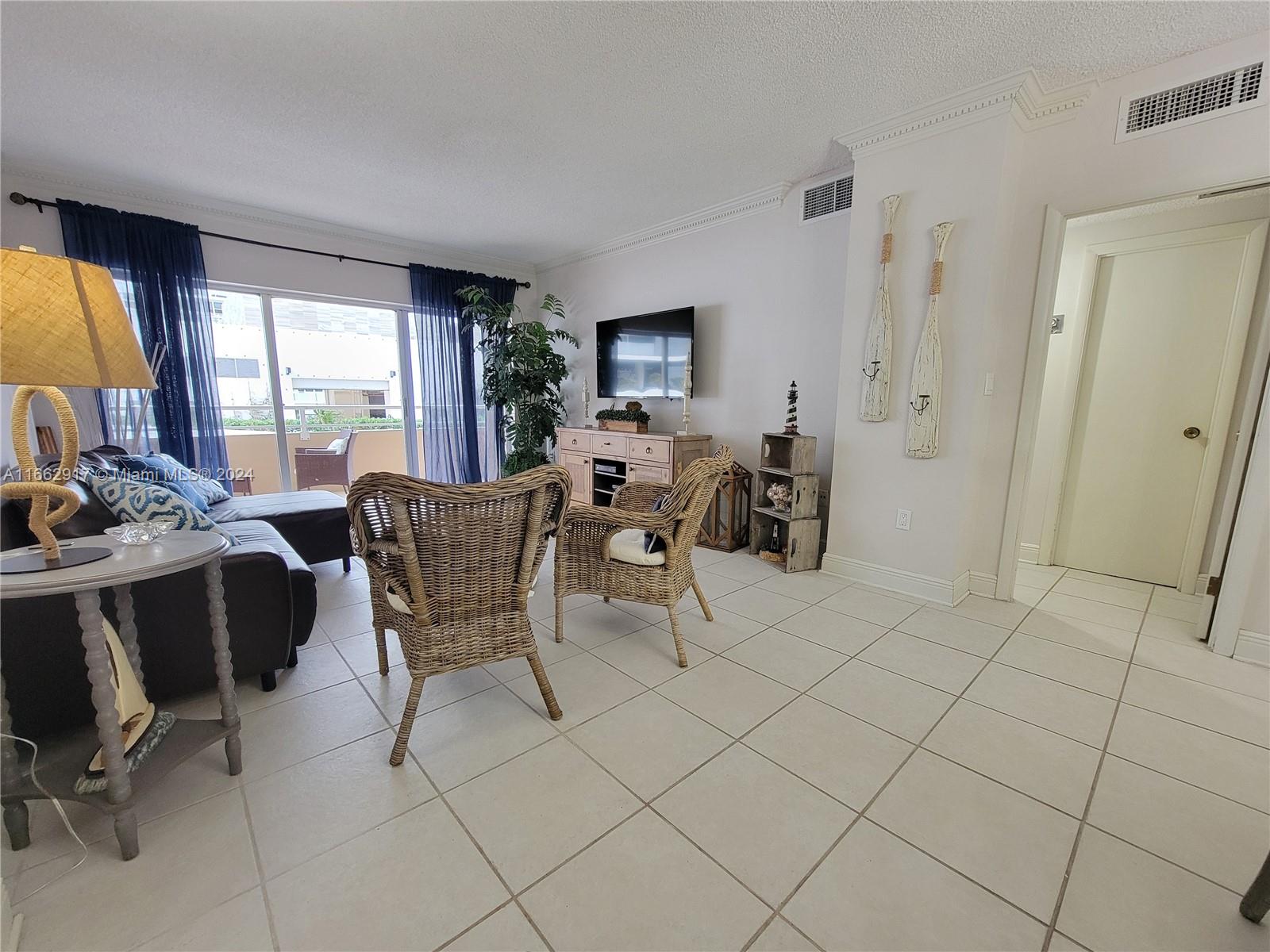 A living room features tile floors, wicker chairs, a television, and a balcony with curtains — 1980 S Ocean Dr # 2C, Hallandale Beach FL 33009, HALLANDALE BEACH, FL