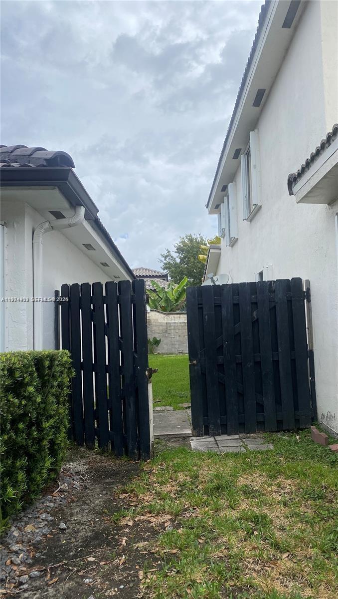 A black wooden gate stands between two white stucco houses leading to a grassy backyard — 87** NW 150th Ter # 0, Miami Lakes FL 33018, MIAMI LAKES, FL