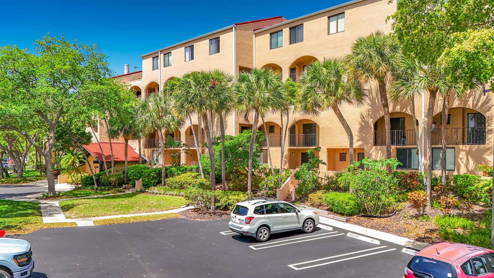 A multi-story tan apartment building with arched balconies sits behind a parking lot with palm trees — 755 Dotterel Road # 1411, Delray Beach FL 33444, DELRAY BEACH, FL