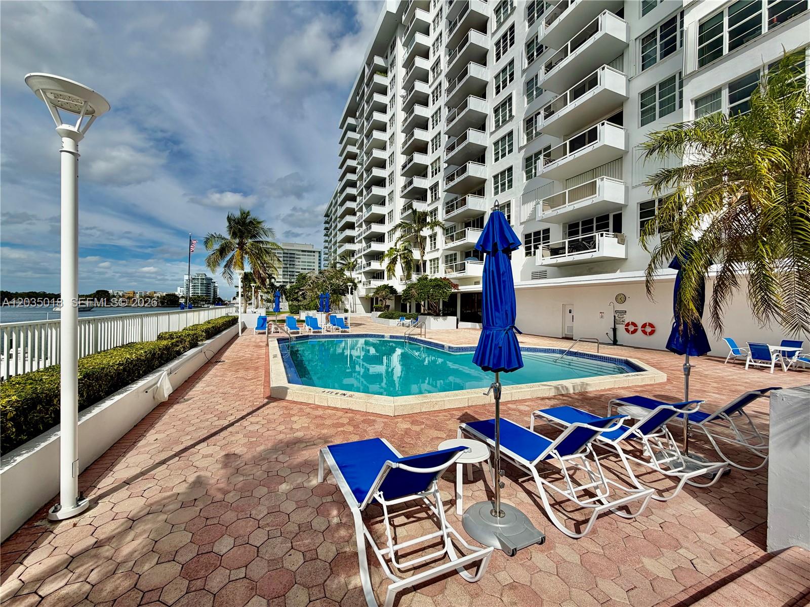 A waterfront patio features lounge chairs, a blue umbrella, and white railings overlooking a canal — 5700 Collins Ave # 14M, Miami Beach FL 33140, MIAMI BEACH, FL