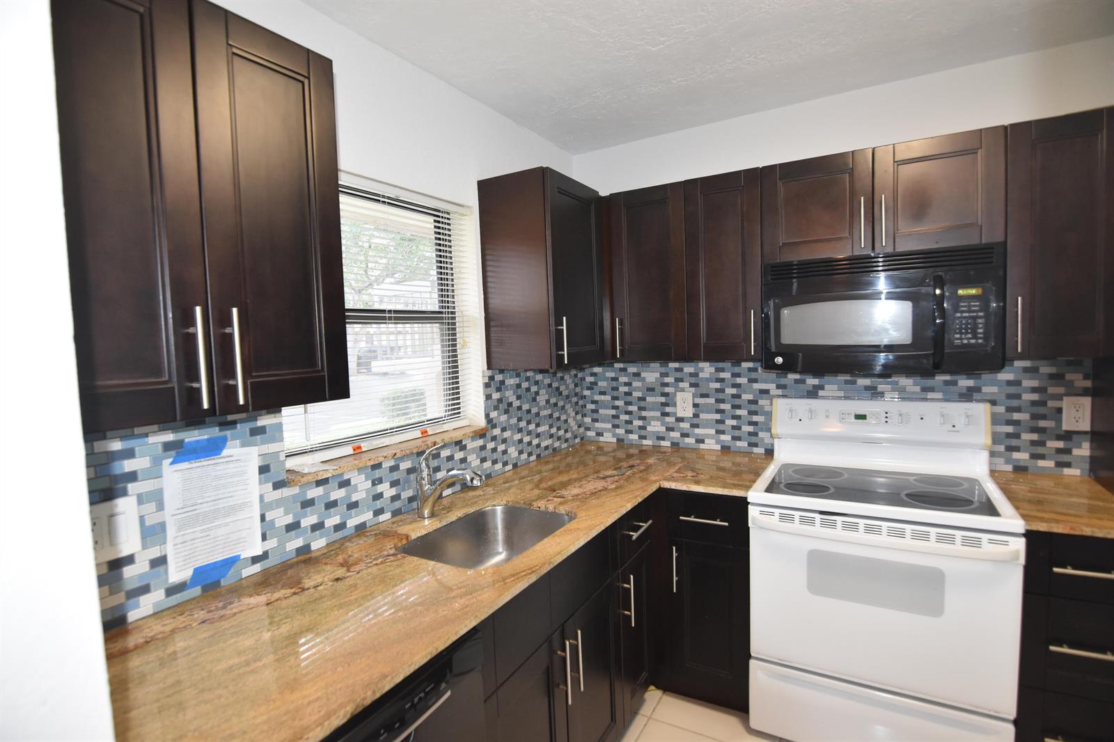A kitchen features dark wood cabinets, granite countertops, a mosaic tile backsplash, and white appliances — 21895 Lake Forest Circle # 102, Boca Raton FL 33433, BOCA RATON, FL