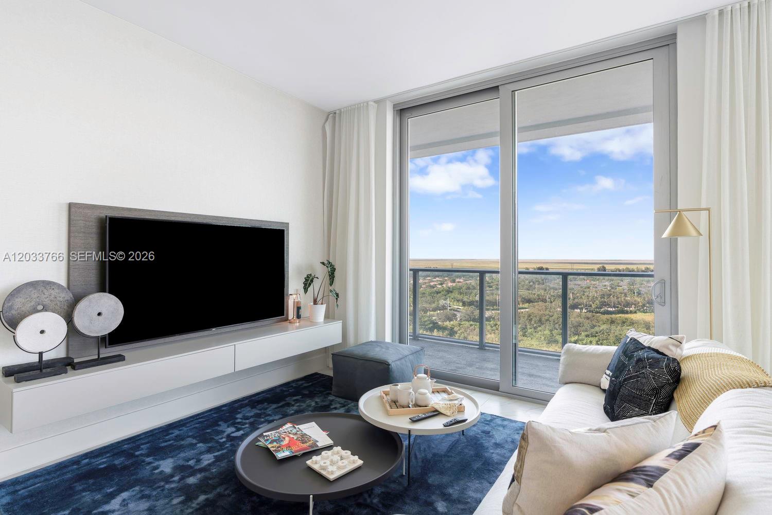 A modern living room features a wall-mounted television, a blue area rug, and a balcony — 2000 Metropica Way # 1810, Sunrise FL 33323, SUNRISE, FL