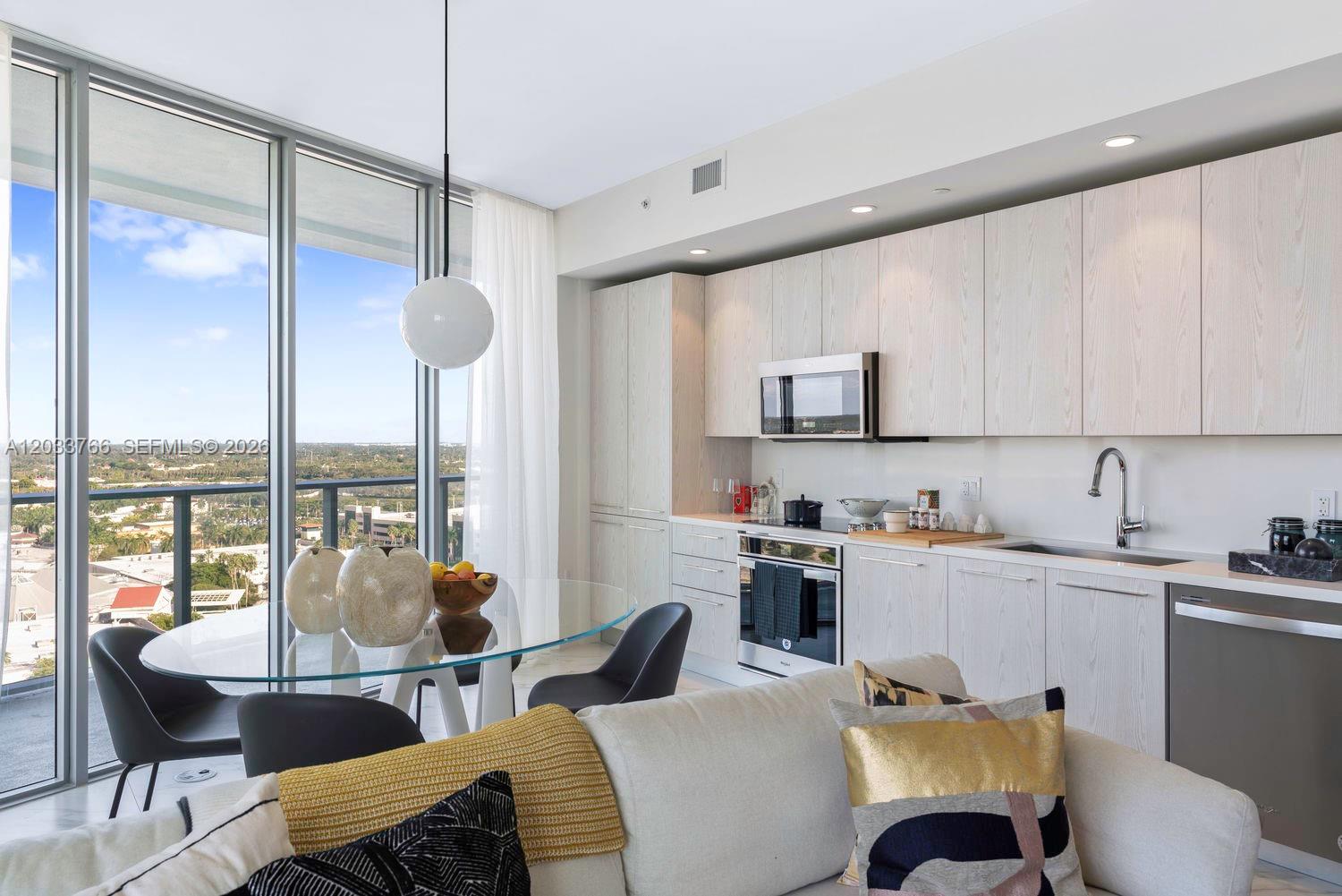 A modern kitchen and dining area feature light wood cabinetry, a glass table, and floor-to-ceiling windows — 2000 Metropica Way # 1810, Sunrise FL 33323, SUNRISE, FL