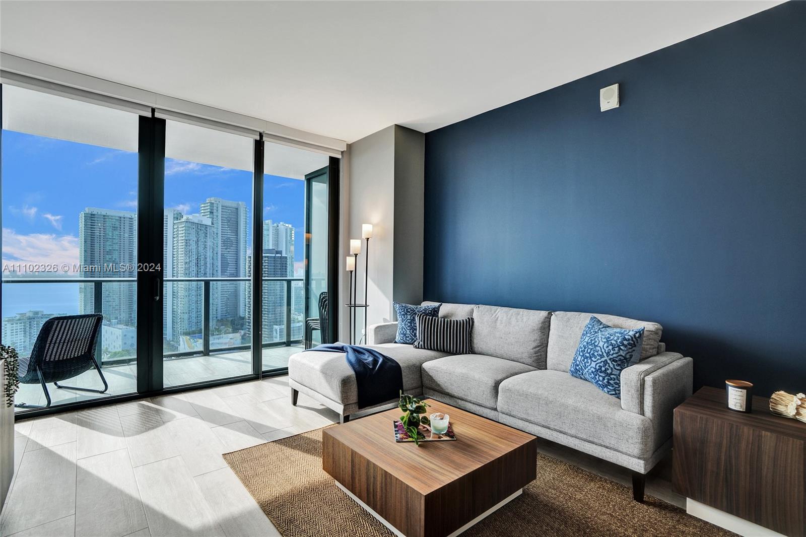 A modern living room features a grey sectional sofa, a wooden coffee table, and floor-to-ceiling windows — 121 NE 34th St # 2906, Miami FL 33137, MIAMI, FL