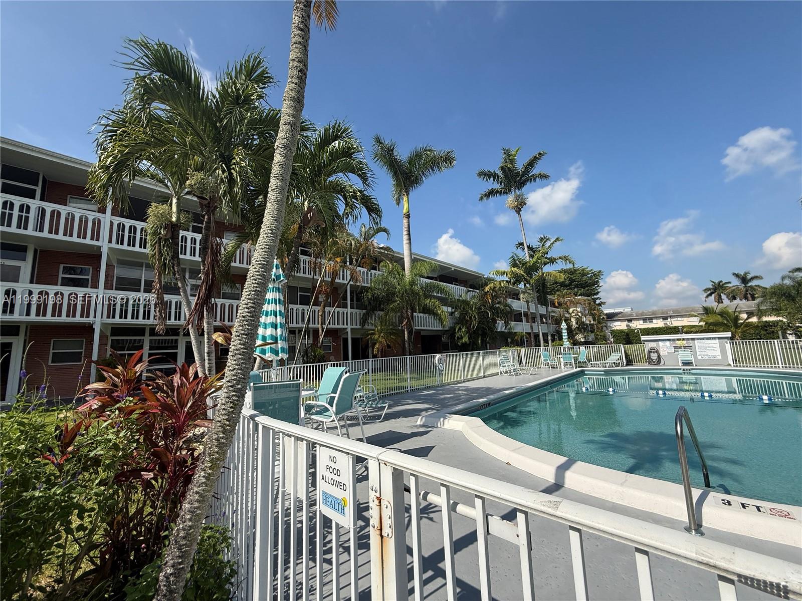 A swimming pool area with lounge chairs, palm trees, and a multi-story apartment building — 141 NE 10th Ave # 17B, Hallandale Beach FL 33009, HALLANDALE BEACH, FL