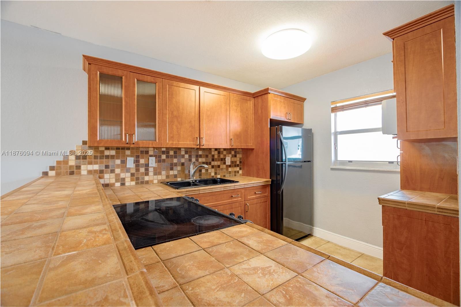 A kitchen features wood cabinets, tiled countertops, a black cooktop, and a stainless steel refrigerator — 120 Royal Palm Rd # 315, Hialeah Gardens FL 33016, HIALEAH GARDENS, FL