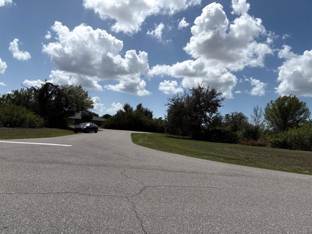 A paved road curves past a grassy lot with trees under a cloudy blue sky — 196 LINDA LEE DRIVE, ROTONDA WEST, FL