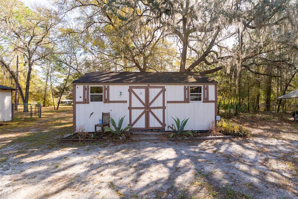 A white storage shed with brown trim and double doors sits in a wooded yard — 14881 NE 147TH AVENUE, FORT MC COY, FL