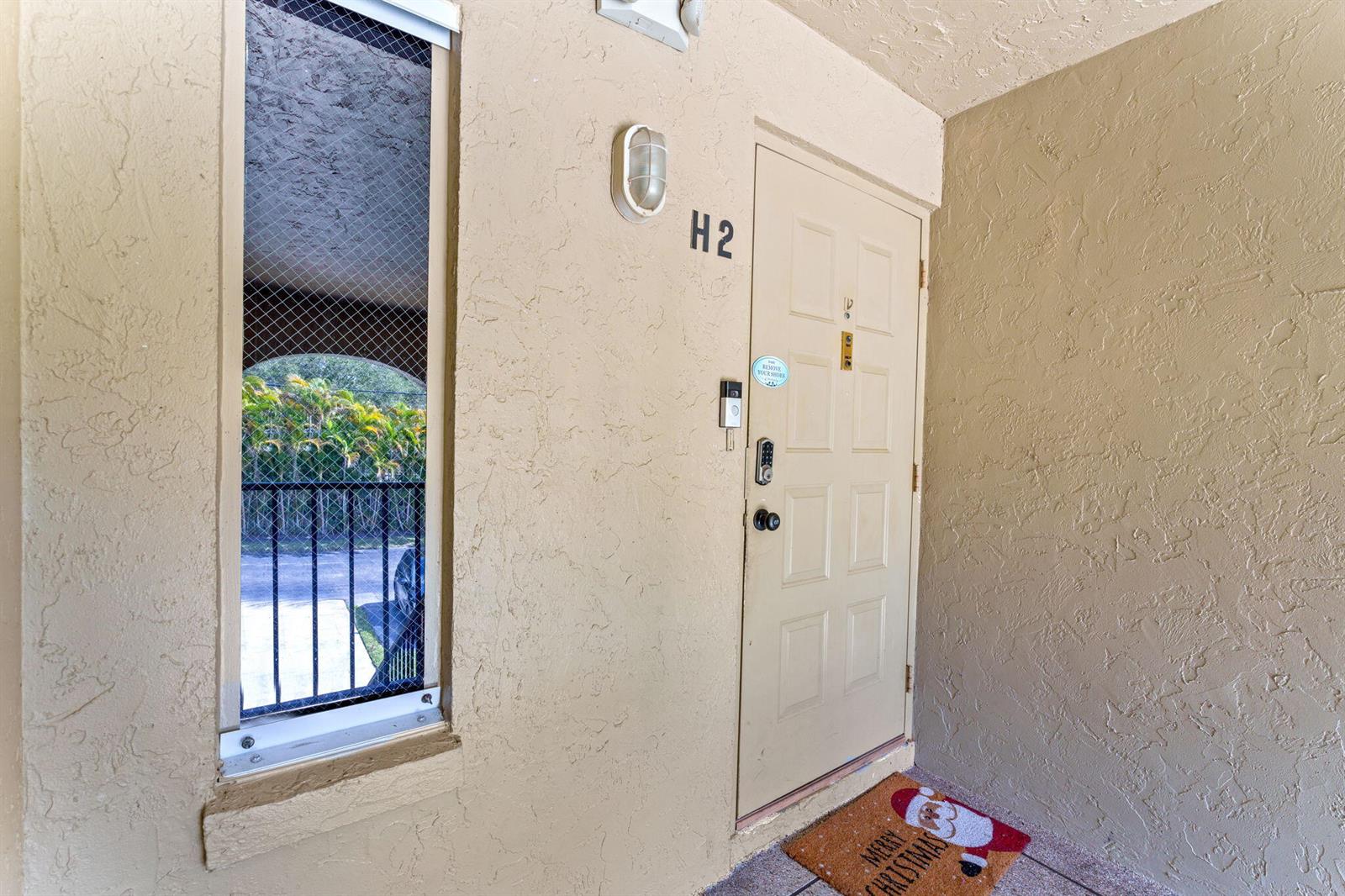 An exterior view of a beige apartment door labeled H2 with a Christmas doormat — 723 Sunny Pine Way # H2, FL 33415, FL