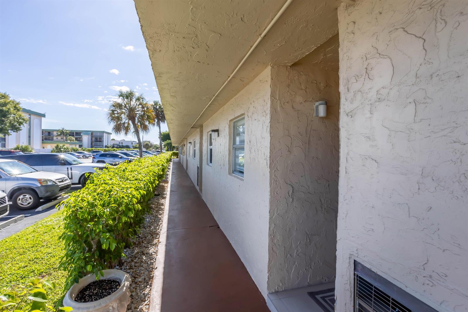 An exterior walkway runs along the side of a stucco building with a hedge — 700 SE 6th Avenue # 107, Deerfield Beach FL 33441, DEERFIELD BEACH, FL