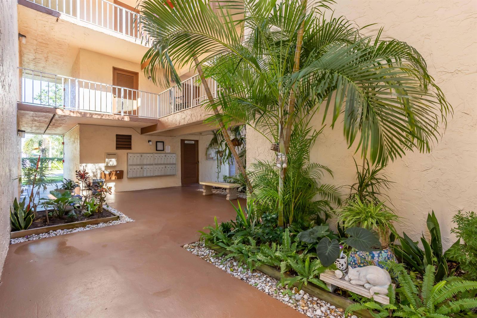 An outdoor courtyard with a palm tree, garden beds, mailboxes, and a second-floor balcony — 700 SE 6th Avenue # 107, Deerfield Beach FL 33441, DEERFIELD BEACH, FL