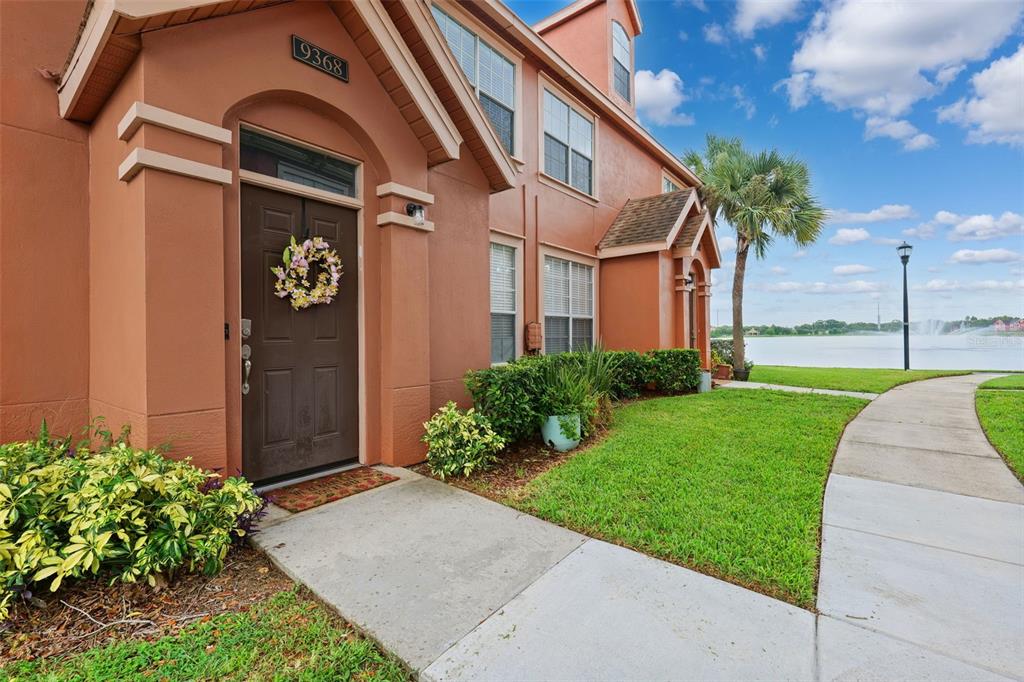 An exterior view of a tan townhouse entrance with a lake and fountain in the background — 9368 LAKE CHASE ISLAND WAY UNIT 9368, TAMPA, FL