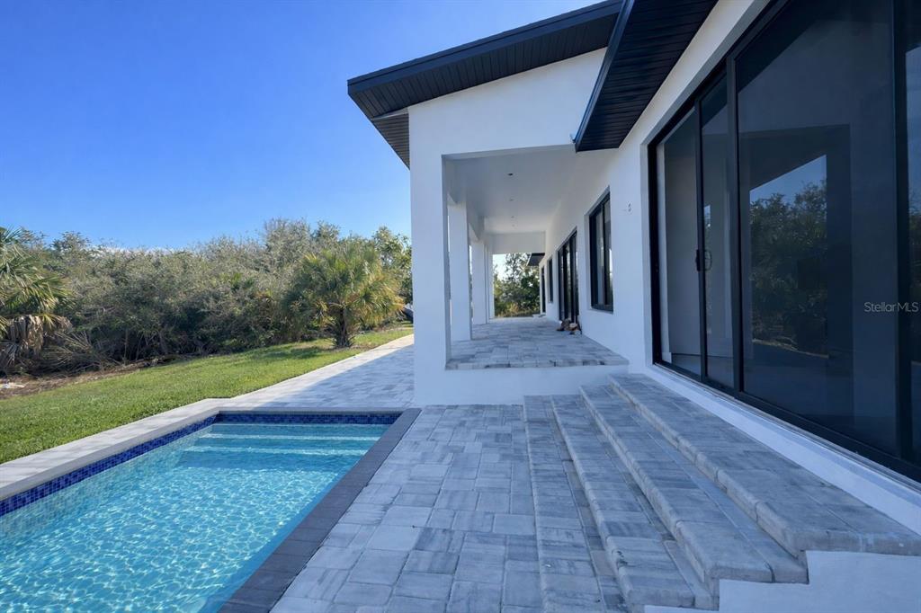 A modern white house exterior features a covered patio, stone pavers, and a rectangular pool — 9477 ATTICA CIRCLE, PORT CHARLOTTE, FL