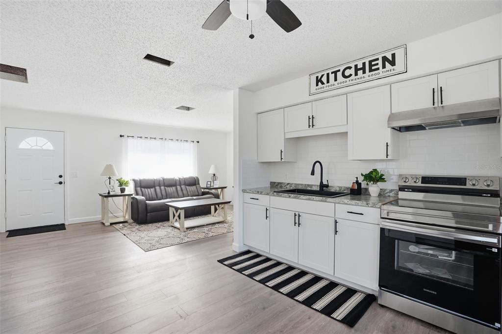 A kitchen with white cabinets and stainless steel appliances opens into a living room area — 2076 LANDOVER BOULEVARD, SPRING HILL, FL