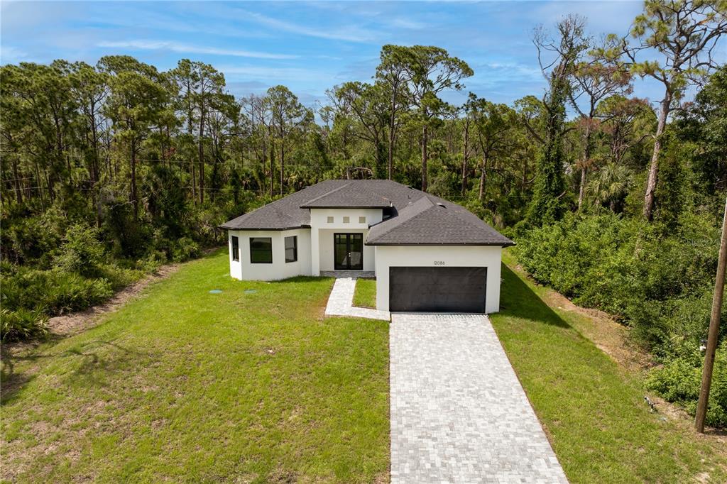 A modern, single-story white house with a dark roof and a paved driveway sits among trees — 12086 CHAMBERLAIN BOULEVARD, PORT CHARLOTTE, FL