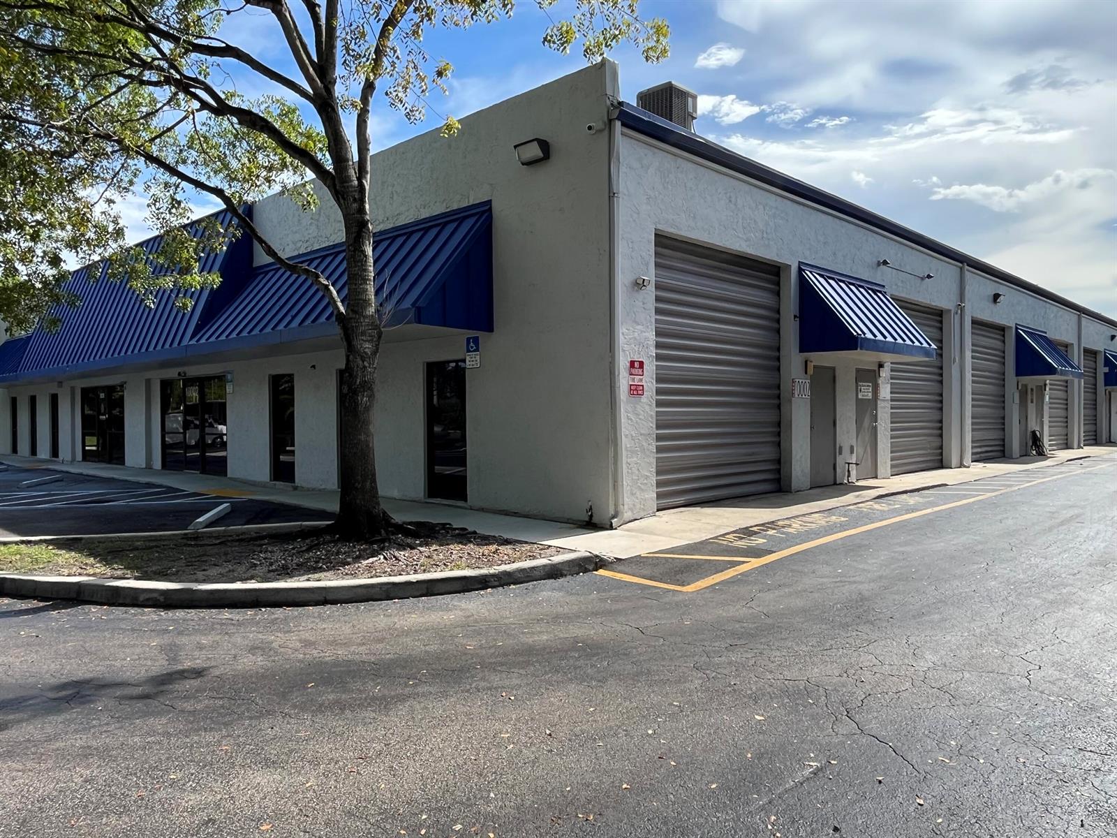 A low-rise commercial building with a white stucco facade, blue metal awnings, and roll-up doors — 10002 NW 46th Street # 10002, Sunrise FL 33351, SUNRISE, FL