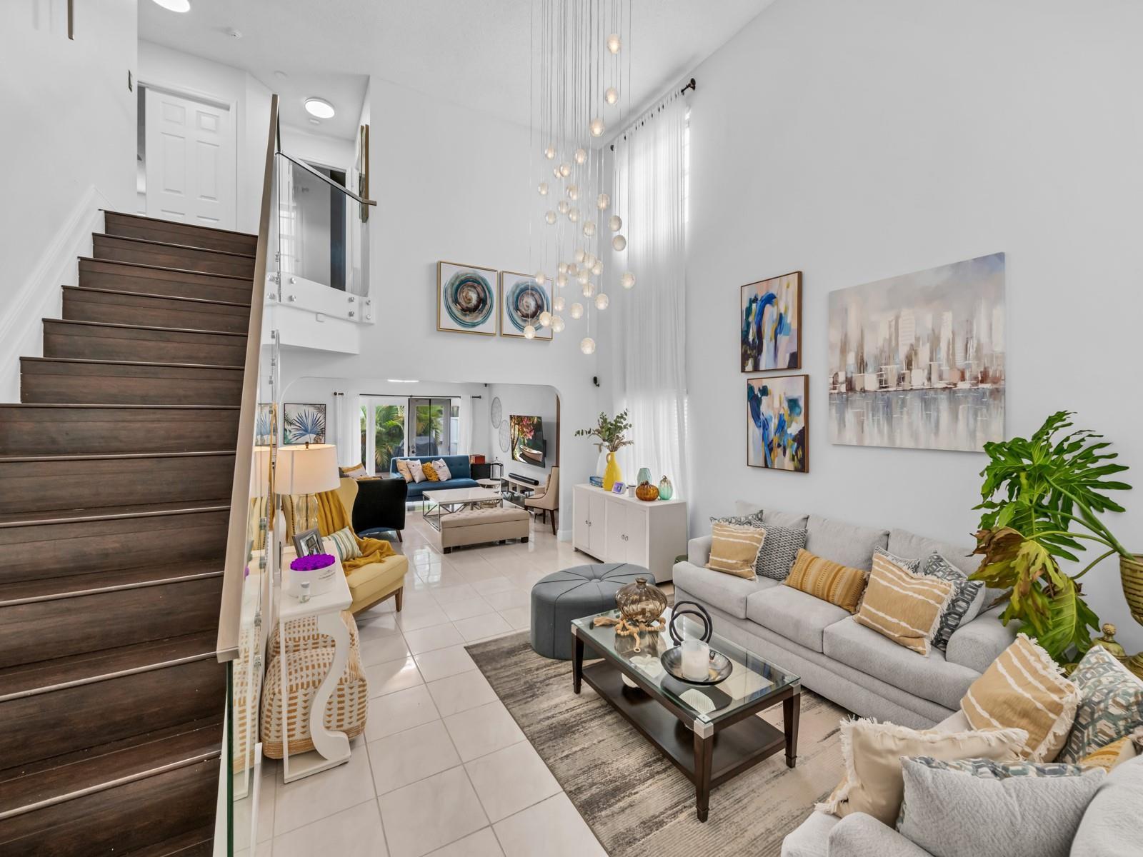 A bright, high-ceilinged living room features a staircase, a modern chandelier, and neutral furniture — 101 Gables Boulevard, Weston FL 33326, WESTON, FL