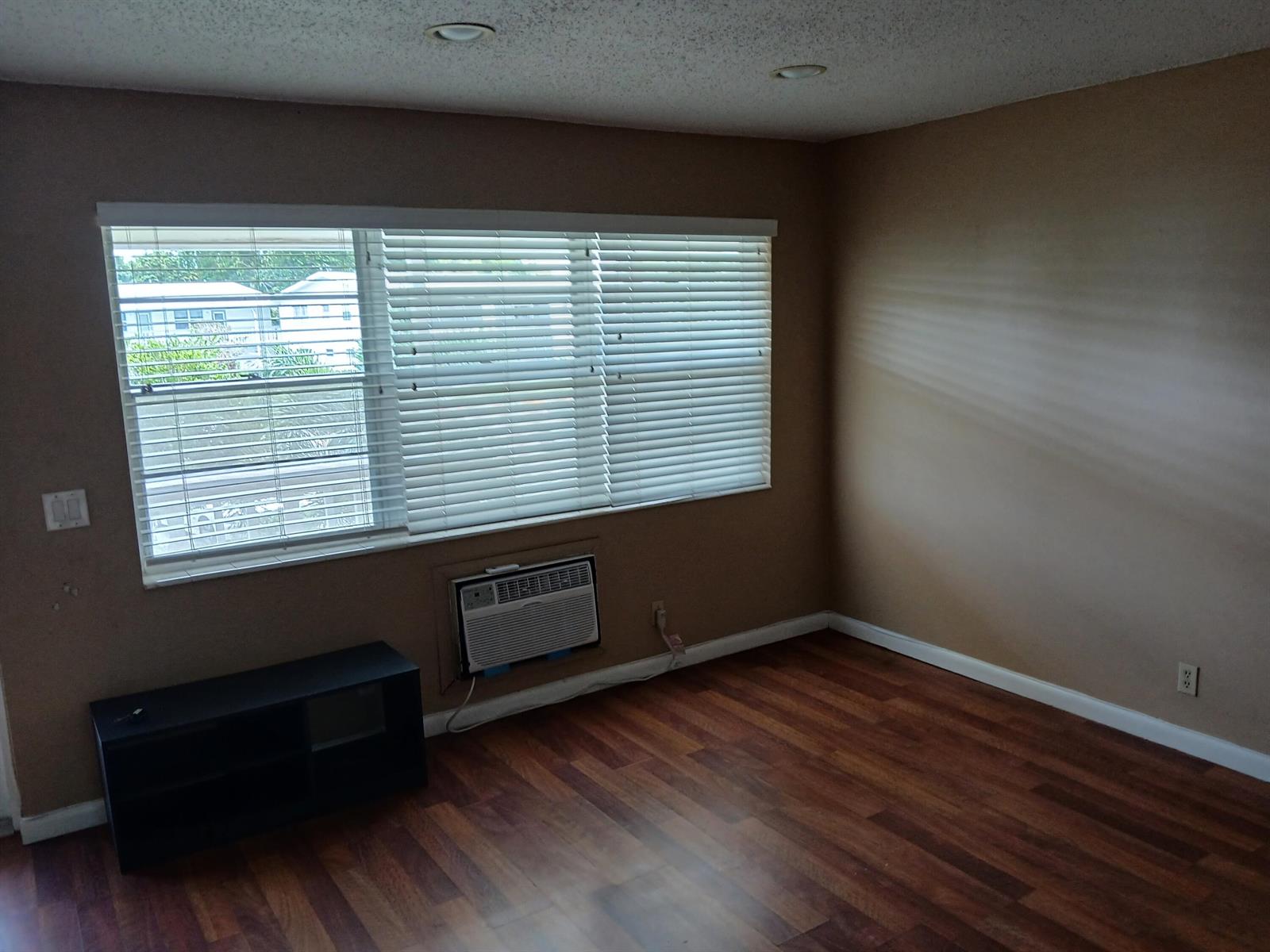 An empty room features brown walls, wood-look flooring, a window with blinds, and an air conditioner — 250 Durham # F, Deerfield Beach FL 33442, DEERFIELD BEACH, FL