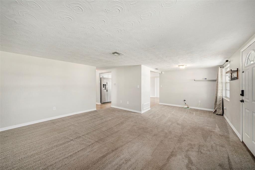 A carpeted living room with light gray walls, a textured ceiling, and an adjacent kitchen — 3510 ALLANDALE DRIVE, HOLIDAY, FL