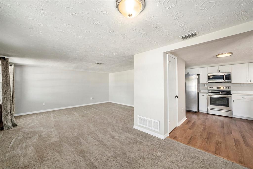 A living room with carpeted floors and a textured ceiling opens into a kitchen area — 3510 ALLANDALE DRIVE, HOLIDAY, FL