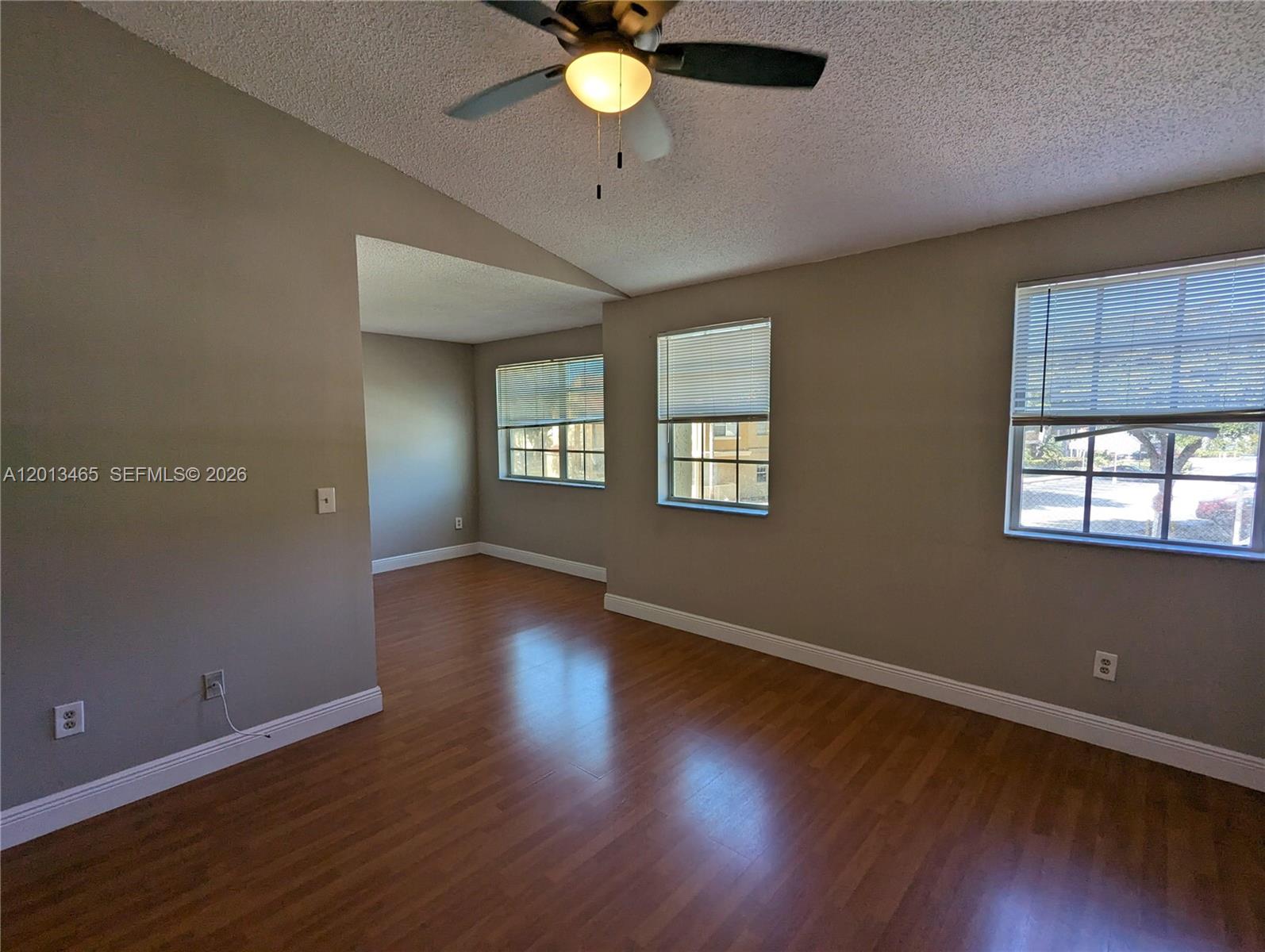 An empty room features wood-look flooring, beige walls, a ceiling fan, and three windows — 18308 NW 68th Ave # D, Hialeah FL 33015, HIALEAH, FL