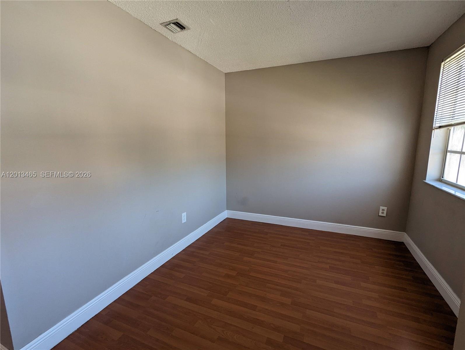 An empty room features beige walls, dark wood-look flooring, white baseboards, and a window — 18308 NW 68th Ave # D, Hialeah FL 33015, HIALEAH, FL