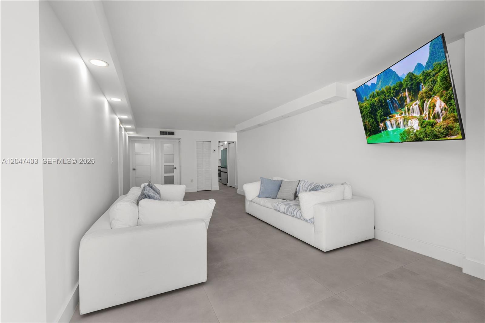 A living room features two white sofas, gray tile flooring, and a wall-mounted television — 2457 Collins Ave # 201, Miami Beach FL 33140, MIAMI BEACH, FL