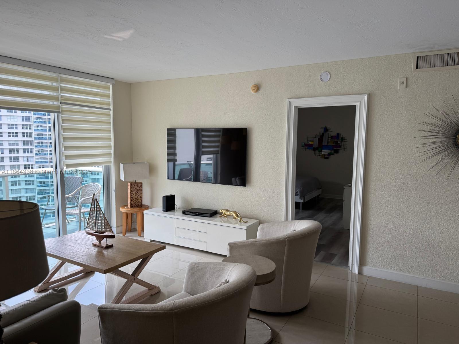 A living room features a wall-mounted television, white media console, two armchairs, and balcony access — 3801 S Ocean Dr # 14V, Hollywood FL 33019, HOLLYWOOD, FL