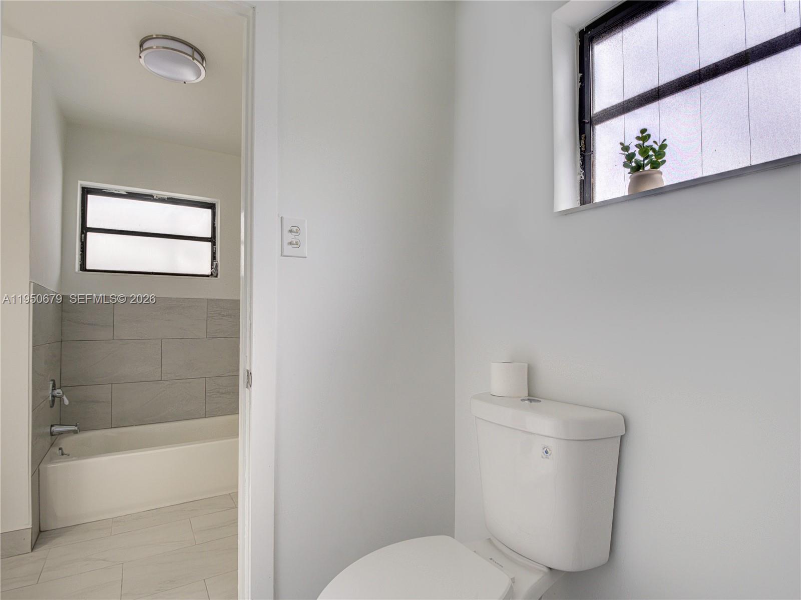 A bathroom features a white toilet in the foreground and a tiled bathtub area beyond — 1500 NW 69th Ter, Miami FL 33147, MIAMI, FL