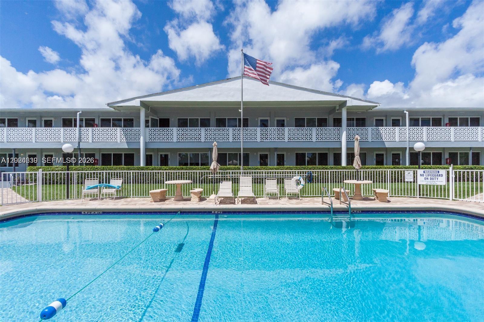 A swimming pool sits in front of a two-story building with a white fence — 600 Layne Blvd # 212, Hallandale Beach FL 33009, HALLANDALE BEACH, FL