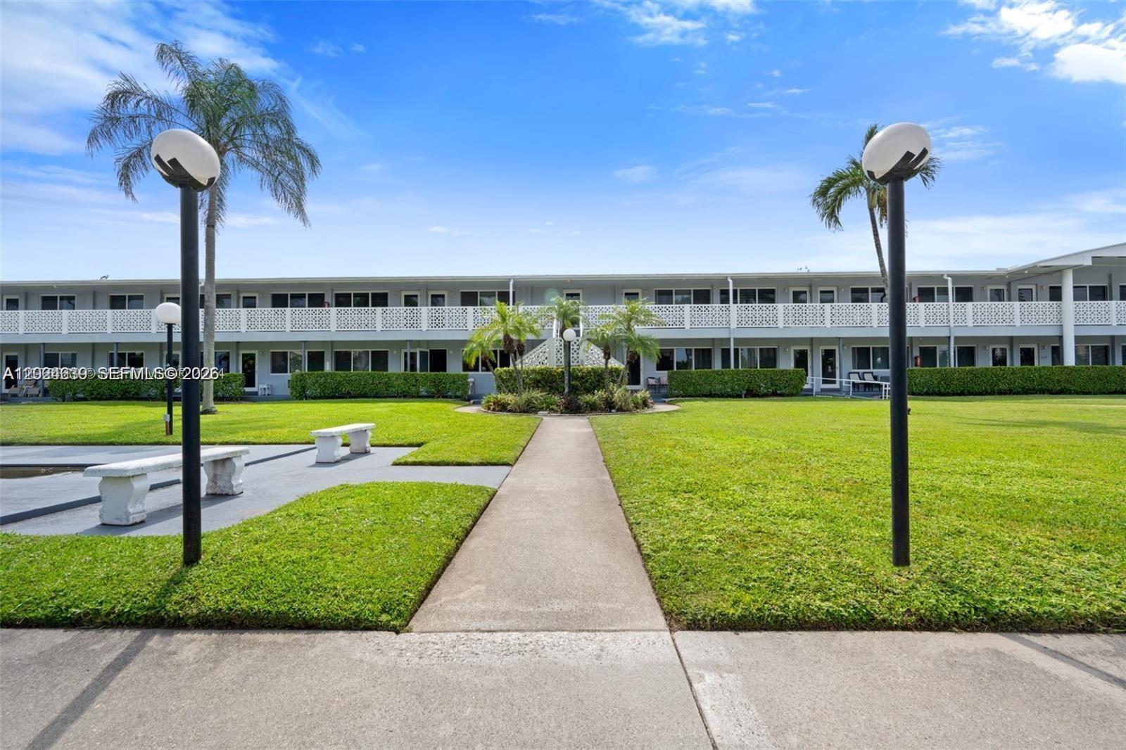 A concrete walkway leads through a grassy courtyard toward a two-story apartment building — 600 Layne Blvd # 212, Hallandale Beach FL 33009, HALLANDALE BEACH, FL