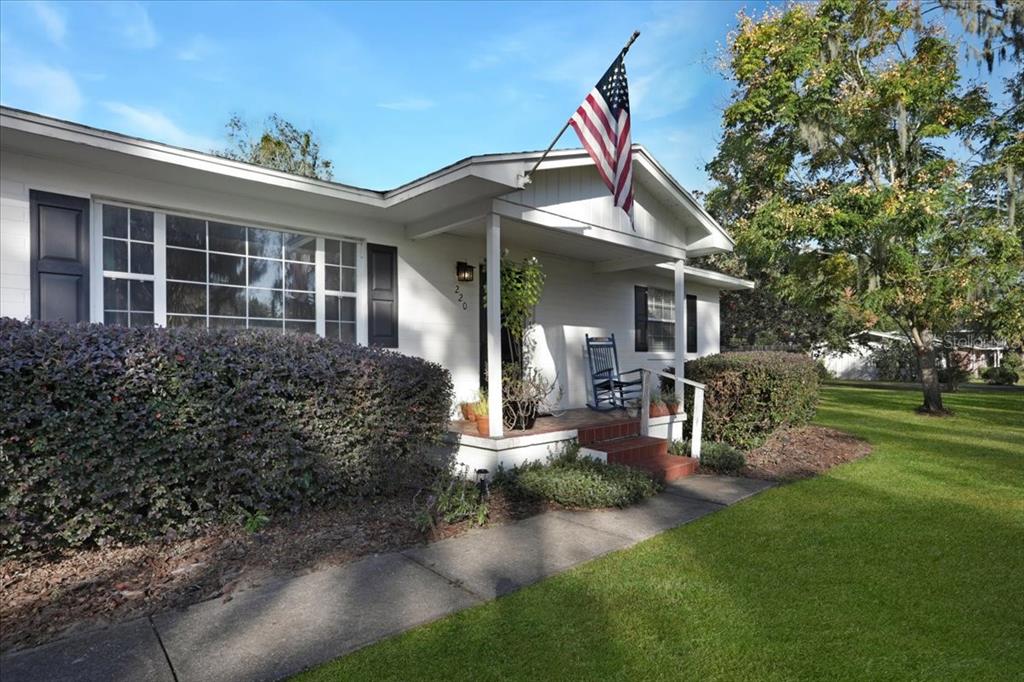 A single-story white house with a front porch, an American flag, and a large hedge — 220 SE 28TH AVENUE, OCALA, FL